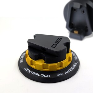 Op de afbeelding: Een zwart-gele Asetek Invicta QR muurbevestiging met een centraal vergrendelingsmechanisme. Het apparaat heeft een zwarte ronde basis met witte tekst die CENTERLOCK, LÖSEN/REMOVE en ANZIEHEN/TIGHTEN leest, naast een zwarte DSS-handgreep.