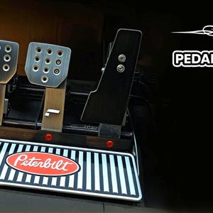 May include: A set of silver and black racing pedals with the Peterbilt logo on the front. The pedals have a textured surface and are mounted on a black base. The logo "Pedal Craft" is visible in the image.