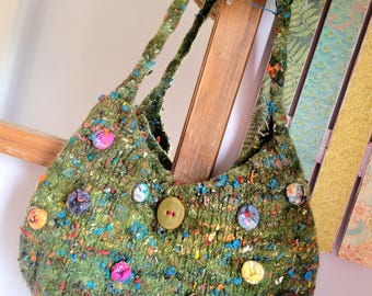 May include: A green, textured handbag with colourful flecks and decorative buttons. The bag has two thick straps and a rounded shape. A straw hat with a black band hangs above the bag, and a wooden ladder is in the background.