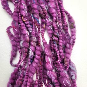 May include: A close-up of a purple yarn with beads woven into the yarn. The yarn is soft and fluffy and has a unique texture.