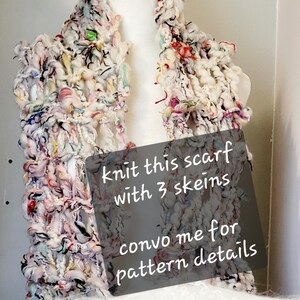 Op de afbeelding: Een witte, roze en zwarte grove gebreide sjaal met kleurrijke accenten. De sjaal wordt getoond op een wit mannequin. De tekst op de afbeelding luidt: "Knit this scarf with 3 skeins convo me for pattern details".