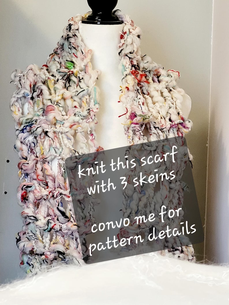 Op de afbeelding: Een witte, roze en zwarte grove gebreide sjaal met kleurrijke accenten. De sjaal wordt getoond op een wit mannequin. De tekst op de afbeelding luidt: "Knit this scarf with 3 skeins convo me for pattern details".