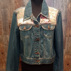 May include: A denim jacket with floral patchwork on the shoulders. The jacket has a classic design with button closures, two chest pockets, and long sleeves. The patchwork features pink and green floral patterns.