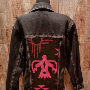 May include: A dark denim jacket with a black back panel featuring a pink Native American-inspired design. The design includes a sun, bird, and geometric shapes. The jacket has a classic collar and button closures.