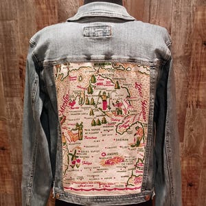 May include: Light blue denim jacket with a detailed embroidered map design on the back. The map features various cities and landmarks in Michigan, with red, green, and tan accents. A stylish and unique outerwear piece.