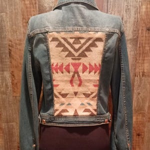 Könnte beinhalten: Jeansjacke mit dekorativem Rückenteil. Das Rückenteil hat ein geometrisches Muster in Beige-, Rot- und Brauntönen. Die Jacke hat einen klassischen Kragen, lange Ärmel und Knopfverschlüsse. Die Jacke wird auf einer Schaufensterpuppe präsentiert.