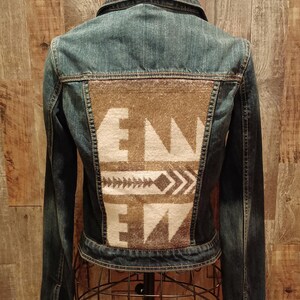 May include: A denim jacket with a brown and white patterned back panel. The back panel features the letters "EN" and geometric designs. The jacket is displayed on a metal dress form.