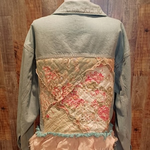 Puede incluir: Una chaqueta vaquera azul claro con un diseño de patchwork en la espalda. El patchwork está hecho de tela floral en tonos de rosa, verde y blanco. La chaqueta tiene un dobladillo con volantes.