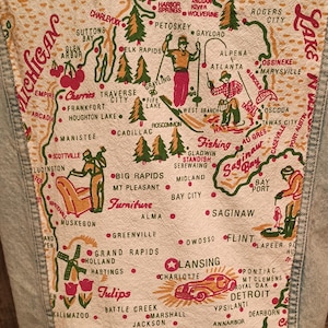May include: Light blue denim jacket with a colorful map of Michigan on the back. The map includes illustrations of skiers, fishermen, and furniture, with city names like Lansing and Detroit. The jacket has a frayed hem.