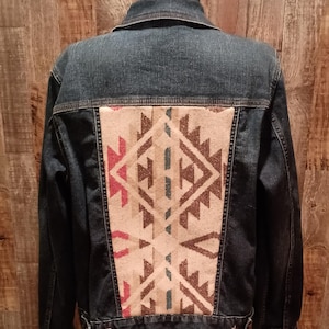 May include: A dark wash denim jacket with a decorative patterned back panel. The panel features a geometric design in shades of beige, brown, red, and blue. The jacket has a classic collar, button closures, and pockets, offering a stylish and unique look.