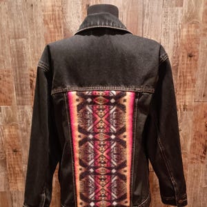 May include: Black denim jacket with a colorful patterned back panel. The panel features a geometric design in shades of red, orange, brown, and cream. The jacket has a classic collar, button closures, and pockets.