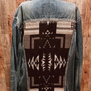 May include: A blue denim jacket with a decorative back panel. The back panel features a geometric pattern in black, gray, and white. The jacket has a classic collar and button closures.