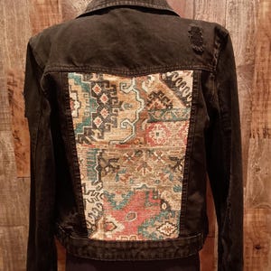 Puede incluir: Chaqueta vaquera negra con un panel de tela estampada en la espalda. El panel de tela presenta una mezcla de colores rojo, turquesa y beige en un diseño geométrico. La chaqueta tiene un cuello clásico y cierre de botones.