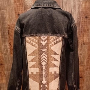 May include: Dark denim jacket with a patterned back panel. The back panel features a geometric design in shades of brown, beige, and white. The jacket has a classic collar, button closures, and long sleeves. The jacket is displayed on a black metal hanger.