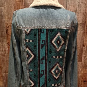 May include: A denim jacket with a cream-colored sherpa collar and a patterned back panel. The back panel features a teal, black, and white geometric design. The jacket has a classic button-up front and buttoned cuffs.