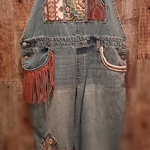 Puede incluir: Mono vaquero azul claro con parches decorativos y flecos. El bolsillo del peto presenta un diseño geométrico colorido. El bolsillo izquierdo tiene un detalle de flecos marrones. Los parches están cosidos en las rodillas. El mono está colgado en una percha rosa.