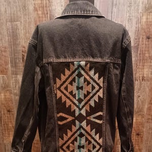 May include: A dark grey denim jacket with a decorative patterned back panel. The back panel features a geometric design in shades of brown, teal, and cream. The jacket has a classic collar and button fastenings.