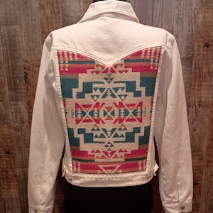 May include: White denim jacket with a colorful geometric patterned back panel. The design features teal, red, and cream-colored shapes. The jacket has a classic collar, button closures, and pockets.