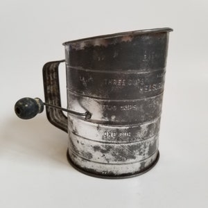 May include: A vintage metal measuring cup with a handle and a rotating knob. The cup is marked with "One Cup", "Two Cups", and "Three Cups".