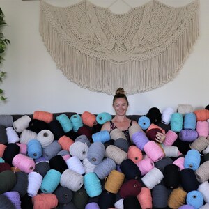 May include: A woman sits surrounded by a variety of colourful cotton rope, likely for macrame projects. The rope is in various shades of pink, blue, green, brown, and white.
