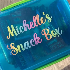 May include: A transparent blue plastic snack box with a green handle. The box is labelled "Michelle's Snack Box" in a colourful, cursive font. Inside, various snack-sized candy packets are visible, including Haribo Supermix.