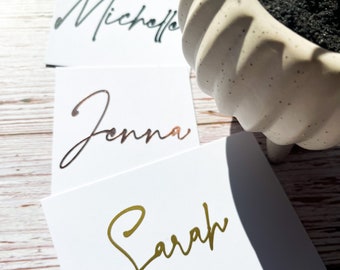 May include: Three white cards with gold, rose gold, and teal calligraphy names: Sarah, Jenna, and Michelle.