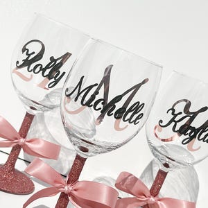 May include: Three clear wine glasses with names in black script and a rose gold accent. The stems are covered in rose gold glitter and tied with a pink satin bow. The names are "Holly", "Michelle", and "Kayla".