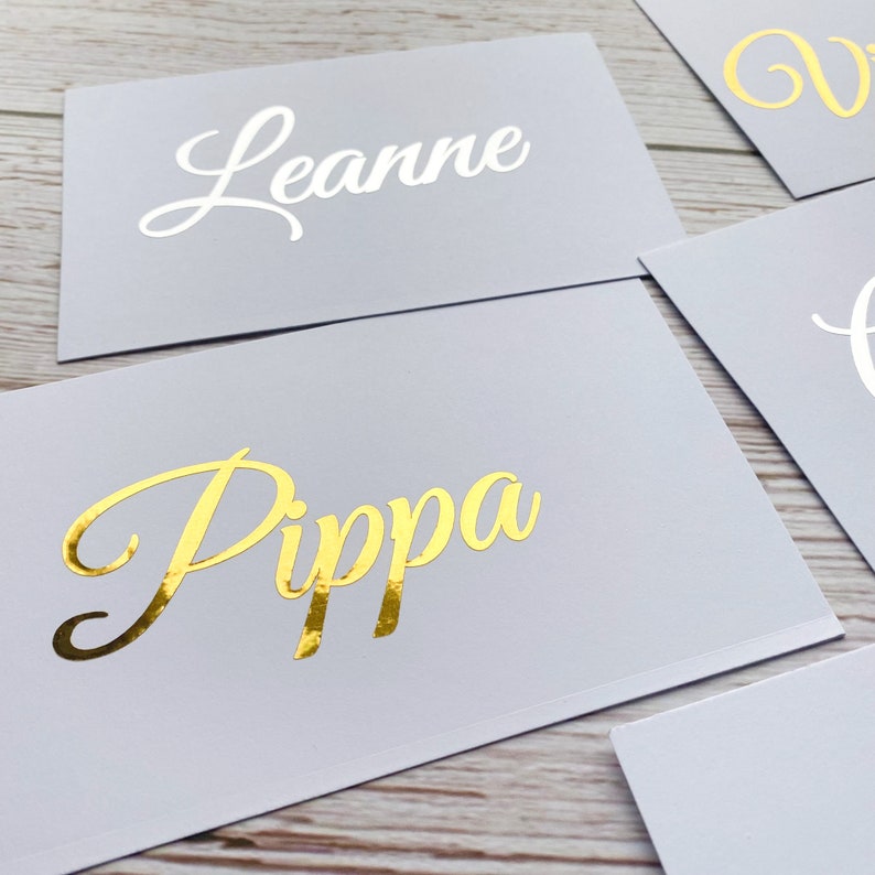 May include: Two white cards with names written in gold and white lettering. The top card says "Leanne" in white lettering and the bottom card says "Pippa" in gold lettering.