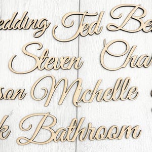 Op de afbeelding: Houten lasergesneden woorden en namen in een cursief lettertype. De woorden zijn onder meer "Wedding", "Ted", "Steven", "Michelle" en "Bathroom". De items zijn gemaakt van lichtgekleurd hout.