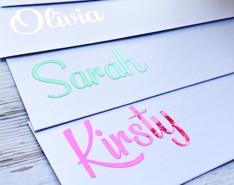 May include: Five white envelopes with different names written on them in colourful foil lettering. The names are Pippa, Victoria, Olivia, Sarah, and Kirsty.