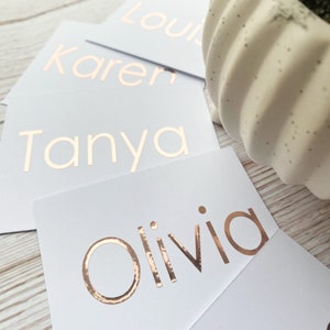 以下が含まれることがあります： 金箔の文字が書かれた白いカードが4枚。カードには、Tanya、Karen、Olivia、Louの名前が書かれています。