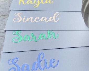 May include: A close-up of several pale blue cards with names in cursive script. The names are in various colors, including yellow, peach, and blue. The names are Kayla, Sinead, Sarah, and Sadie. A white, textured object is in the background.