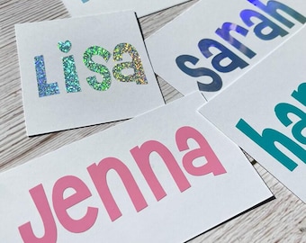 May include: A selection of white rectangular cards, each bearing a name in a bold font. The names are in a variety of colours, including pink, teal, and glitter. The cards are arranged on a light wooden surface.