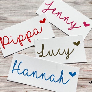 Puede incluir: Cuatro tarjetas blancas con diferentes nombres escritos en cursiva de colores. Los nombres son "Jenny", "Pippa", "Lucy" y "Hannah". Cada nombre tiene un pequeño corazón al lado en un color que coincide con el nombre.