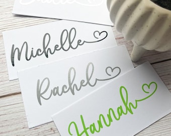 May include: White rectangular cards with names in cursive script, including "Sadie," "Michelle," "Rachel," and "Hannah." The names are in various colours: white, silver, and green. Each name is followed by a heart symbol. A white ceramic pot is in the background.