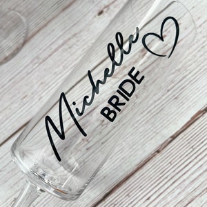 May include: A clear glass champagne flute with a black vinyl sticker that says "Michelle Bride" with a heart design.