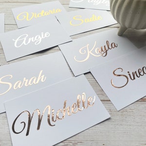 May include: A set of place cards with names written in gold and white lettering on a light grey background. The names include Angie, Sarah, Victoria, Sadie, Kayla, Michelle, and Sineo.