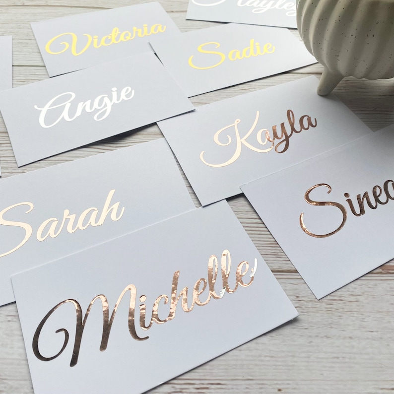 May include: A set of place cards with names written in gold and white lettering on a light grey background. The names include Angie, Sarah, Victoria, Sadie, Kayla, Michelle, and Sineo.