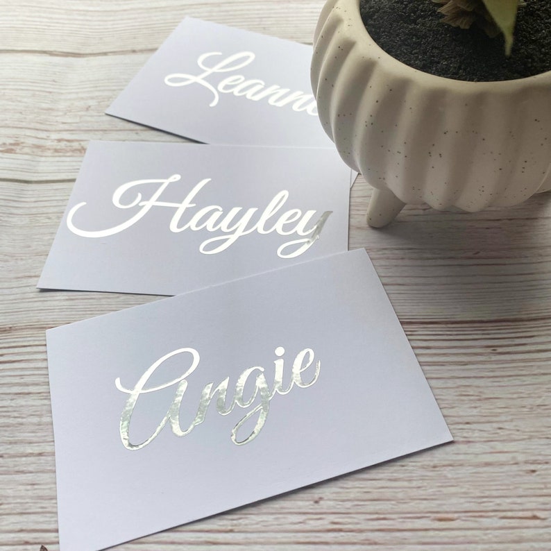 May include: Three white cards with silver foil lettering. The names Hayley, Angie, and Leanne are written on the cards.