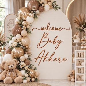 May include: An arched backdrop in cream featuring the brown text "welcome Baby Akhere" decorated with a balloon garland in shades of brown, gold, and white, accented with greenery and flowers, plus a large teddy bear and a "BABY" block tower.