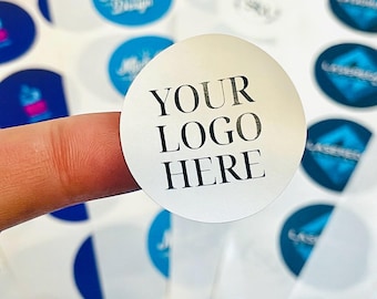 May include: A white, round sticker with the text "YOUR LOGO HERE" in black. The sticker is held by a finger, with other stickers visible in the background. The background stickers have various logos and designs.