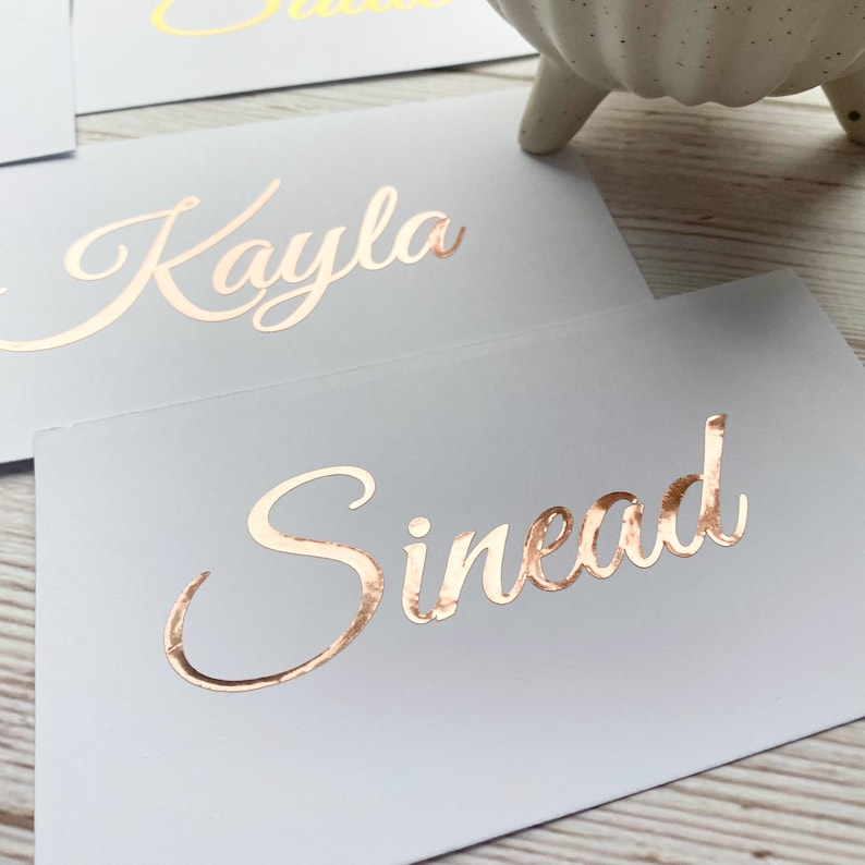 May include: Two white cards with the names "Kayla" and "Sinead" written in gold foil lettering.
