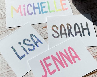 May include: Four white cards with different names written on them in colorful, glittery, and holographic lettering. The names are "Michelle", "Sarah", "Lisa", and "Jenna".