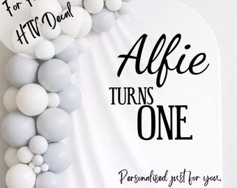 May include: A white background with a balloon arch in shades of white and gray. The text reads "Alfie Turns One" in a stylish font. Additional text includes "For Fabrics HTV Decal" and "Personalised just for you. CHOICE OF SIZE, COLOUR & FONTS."