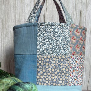 May include: A patchwork tote bag in shades of blue, featuring floral and textured fabrics. The bag has a light blue base and gingham patterned handles. A skein of green yarn is in the foreground.