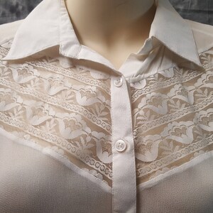 May include: A white long-sleeved button-down shirt with a lace overlay on the front. The lace is a delicate pattern with a floral design.