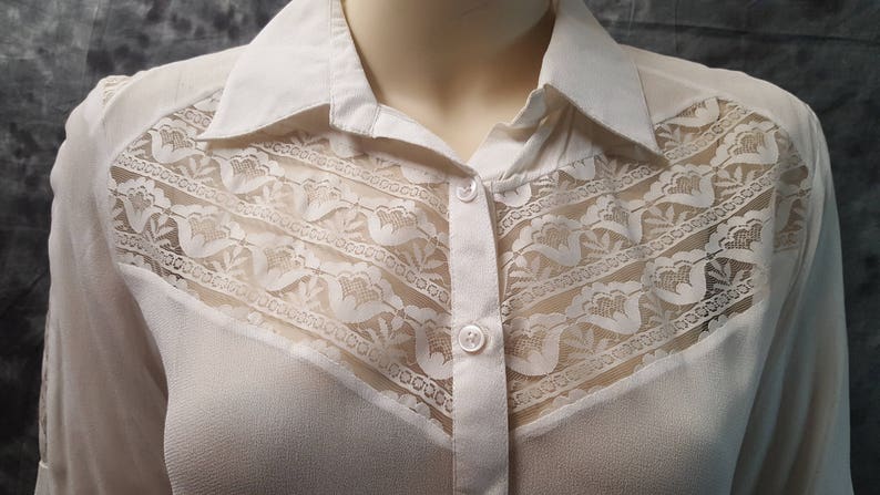 May include: A white long-sleeved button-down shirt with a lace overlay on the front. The lace is a delicate pattern with a floral design.