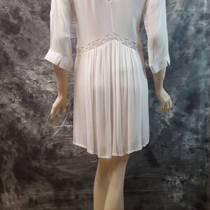 May include: White, long-sleeved dress with a lace overlay on the back. The dress has a collared neckline and a pleated skirt.