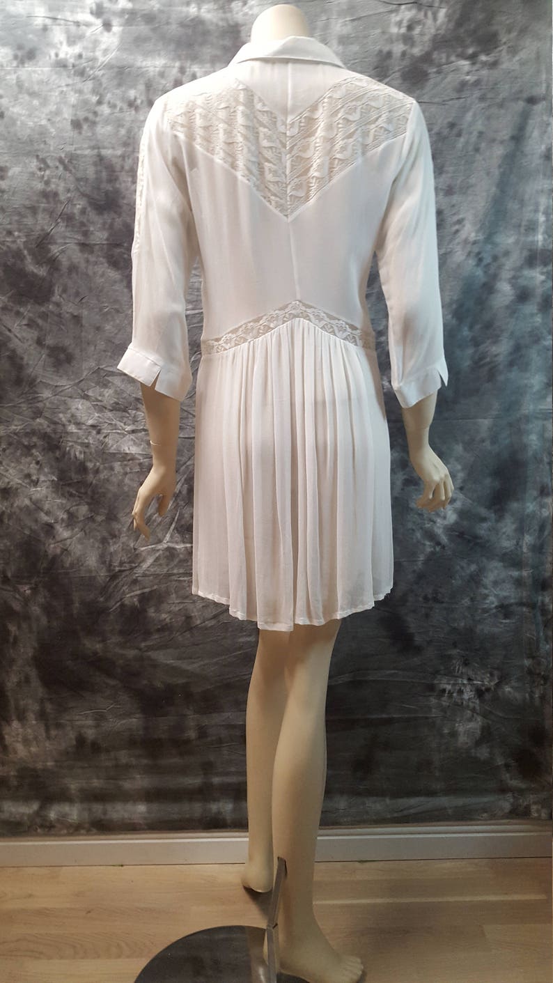 May include: White, long-sleeved dress with a lace overlay on the back. The dress has a collared neckline and a pleated skirt.