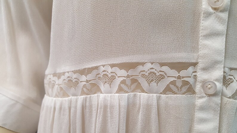 May include: White button-up shirt with a lace trim and a pleated skirt. The shirt has two buttons on the front.